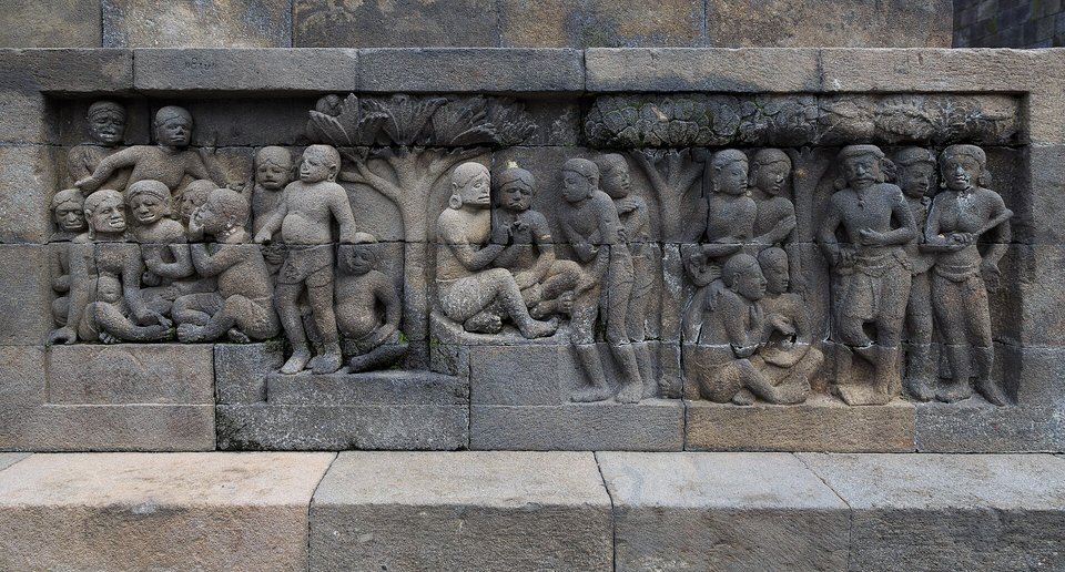 A long stone panel carved in low relief with many robed figures, some seated beneath a tree, framed by plain stone blocks