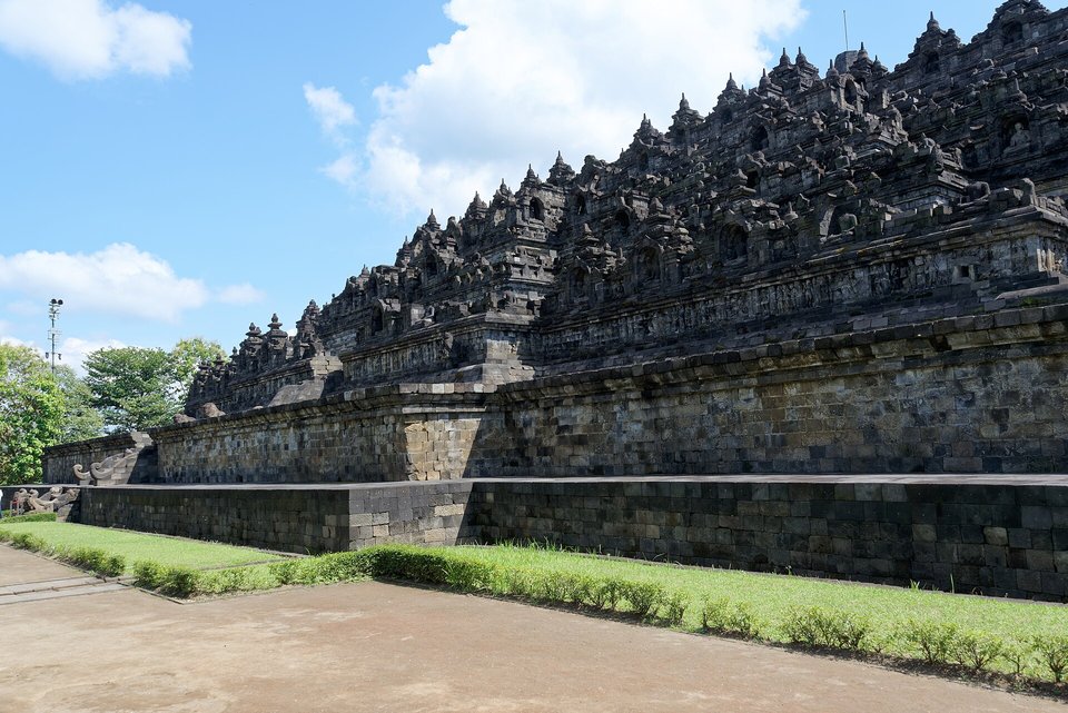 A long dark stone wall of stepped terraces crowded with small stupas and shrines, rising above a grassy path under a blue sky with white clouds