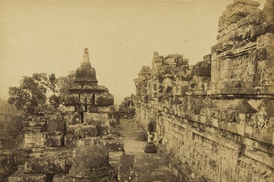 A sepia photograph of a narrow stone-paved gallery between a wall carved with reliefs and a row of stupas, a man seated in dark clothes beside the wall