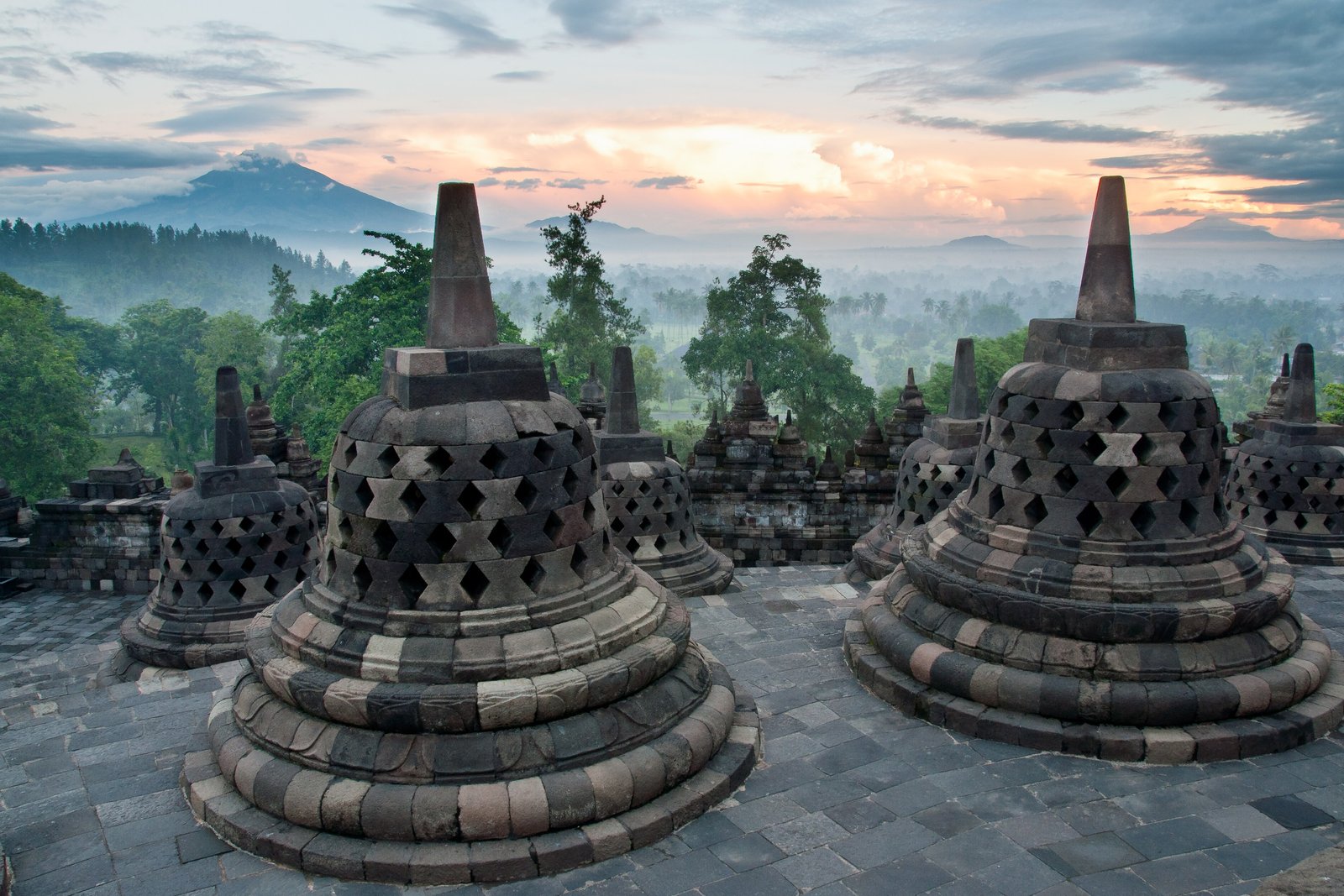 Bell-shaped stone stupas pierced with diamond-shaped openings on a paved terrace, a misty green plain and distant mountains beneath a pink and grey dawn sky