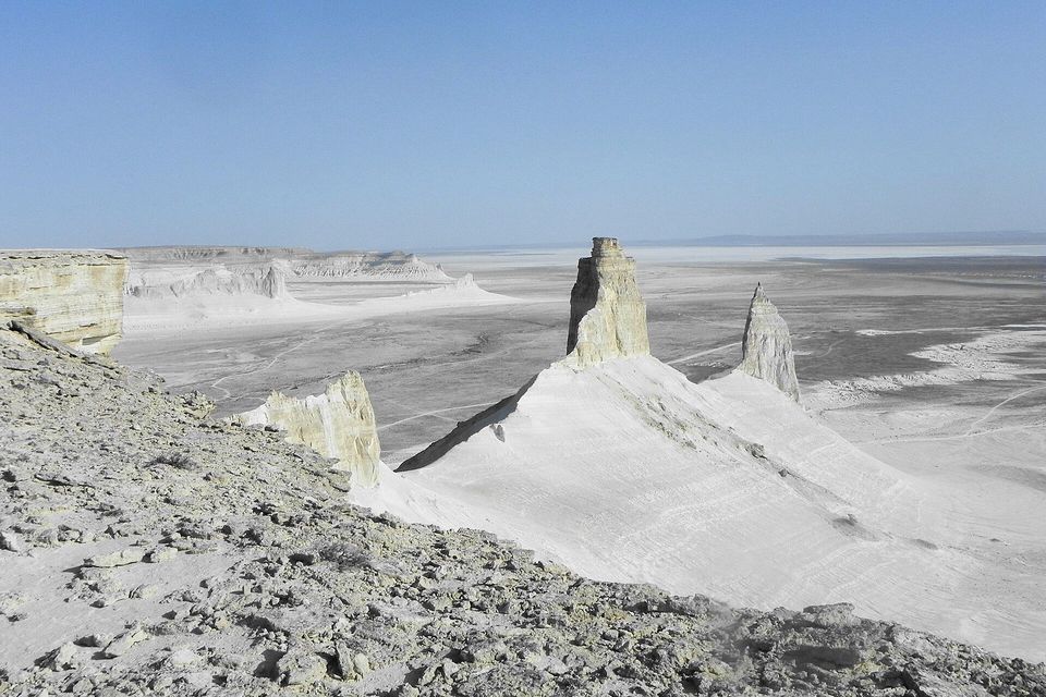 Two pale chalk towers rise from a smooth white ridge over a flat grey-white basin stretching to the horizon, a cliff edge at left