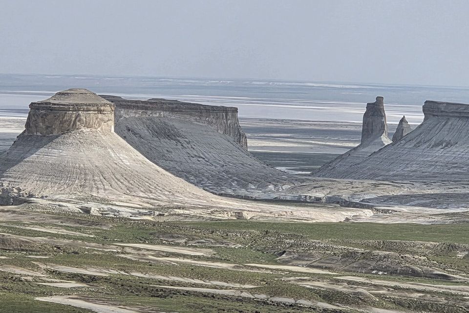 Flat-topped white mesas with ribbed scree skirts stand above a green-grey plain, more towers and a pale basin in haze behind