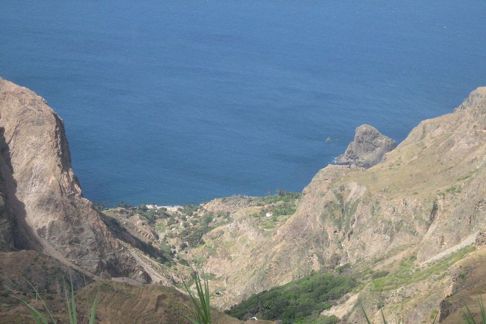 A steep valley of terraced slopes and green scrub drops between brown ridges to a deep-blue sea, a rock stack standing offshore