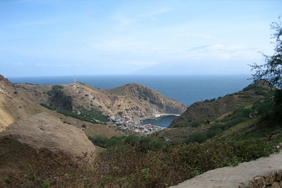 From a stone wall on a dry hillside, a small harbour town sits in a narrow bay below brown hills, the sea and a faint island silhouette beyond