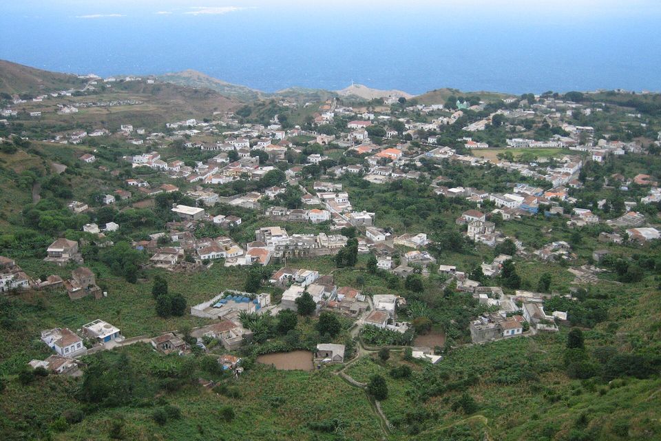 Seen from a high slope, a spread-out town of white and orange-roofed houses sits among green fields and trees, the ocean beyond