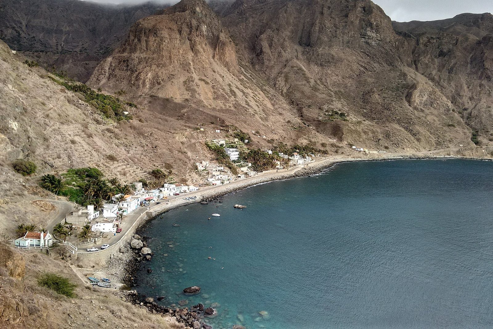 From the cliff road, a curve of teal bay under brown volcanic peaks, the white village of Fajã d'Água strung along the sea wall among palms, cloud resting on the summits