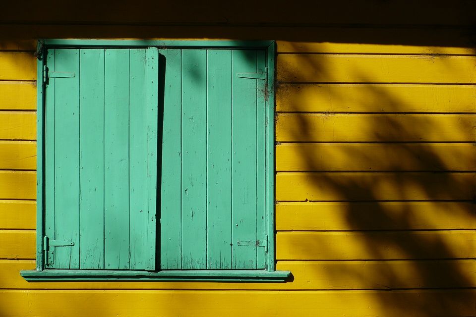 A yellow clapboard wall with a pair of green wooden shutters, leaf shadows falling across it in hard sun