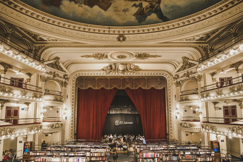 A theatre interior of stalls, a stage and boxes under a painted ceiling, now lined with bookshelves, a few readers small in the stalls