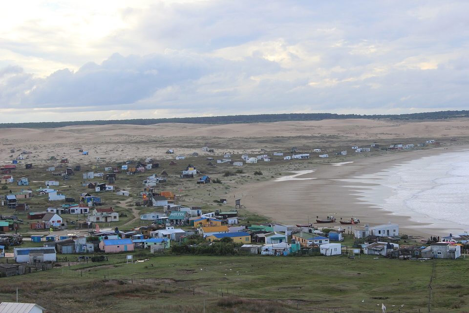 Houses scattered over grass beside a curving beach, with bare dunes stretching away to a tree line