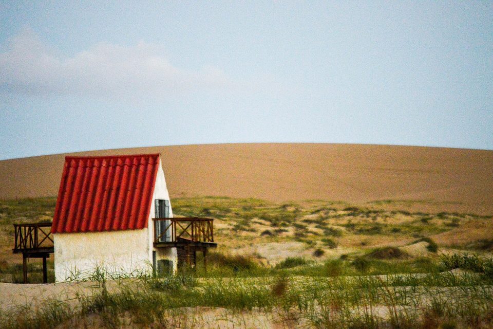 A white house with a red roof below bare sand dunes