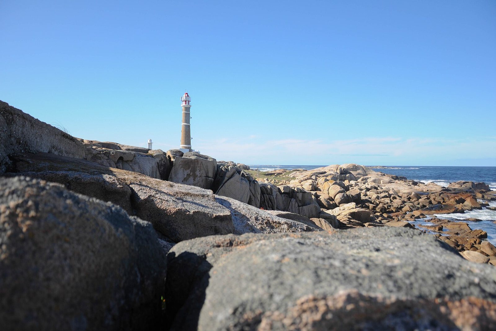 A lighthouse on a cape beyond rounded granite boulders, with surf and open sea to the right under a clean blue sky