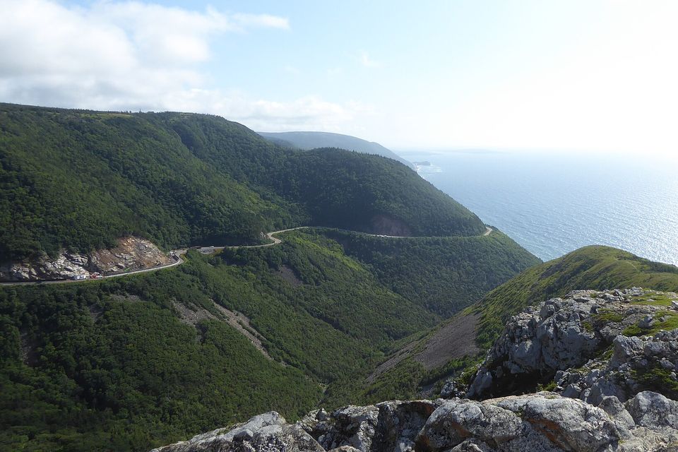 The road looping down a forested mountainside toward the sea, grey rocks in the foreground and the open water glittering at right