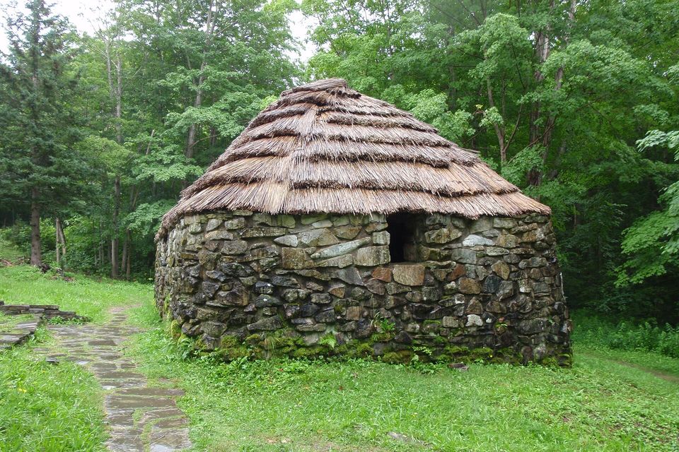 A low round hut of mossy stacked stones under a thick thatched roof, a flagstone path leading past it through green grass, tall hardwood trees behind