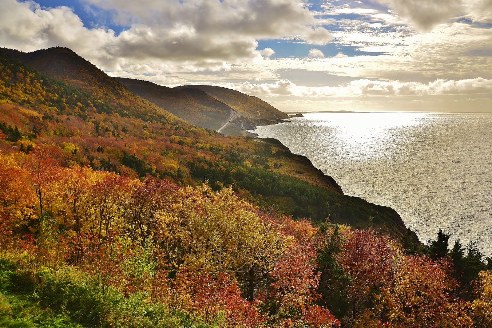 The Cabot Trail cut into the flank of a mountain above the sea, slopes in a blaze of red, orange and gold autumn trees in the foreground, sun glittering on the Gulf of St. Lawrence under broken cloud