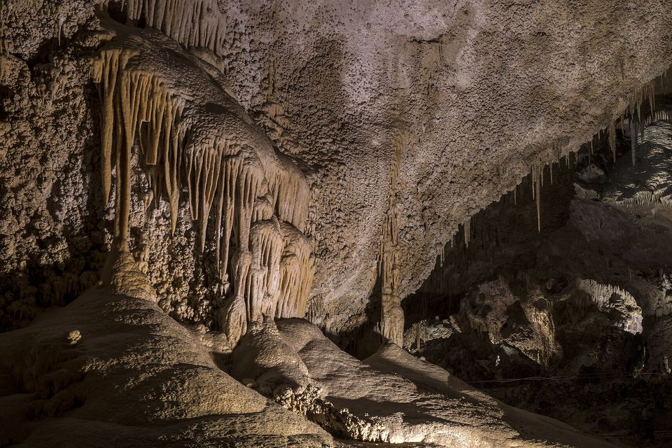 Rippled curtains of pale flowstone pour down a cave wall onto a smooth mound, the ceiling textured like popcorn, a lit chamber beyond at right