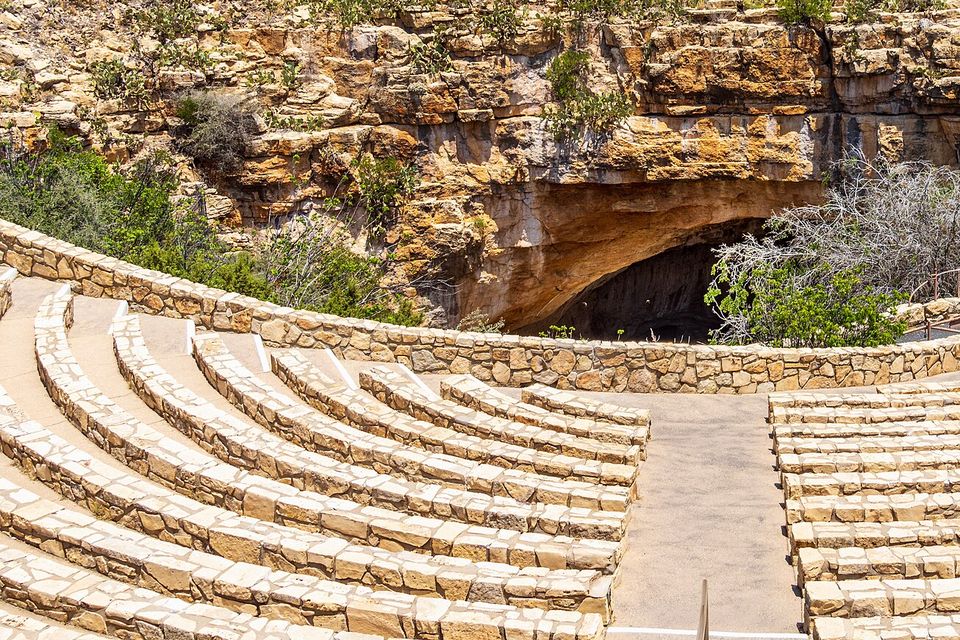 Curved stone benches of an outdoor amphitheatre face a dark cave mouth in a limestone cliff, desert scrub and yucca on the hillside above under a blue sky