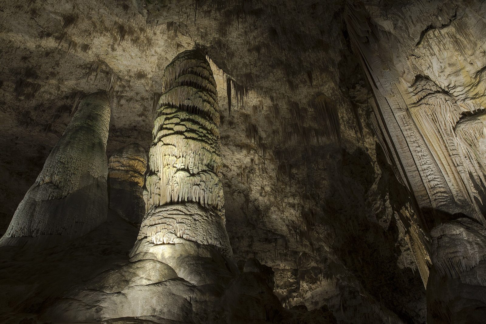 Three giant stalagmite columns lit warm from below rise into a ceiling of soda straws, the middle one ringed in shelves of flowstone, in a vast dark cave room