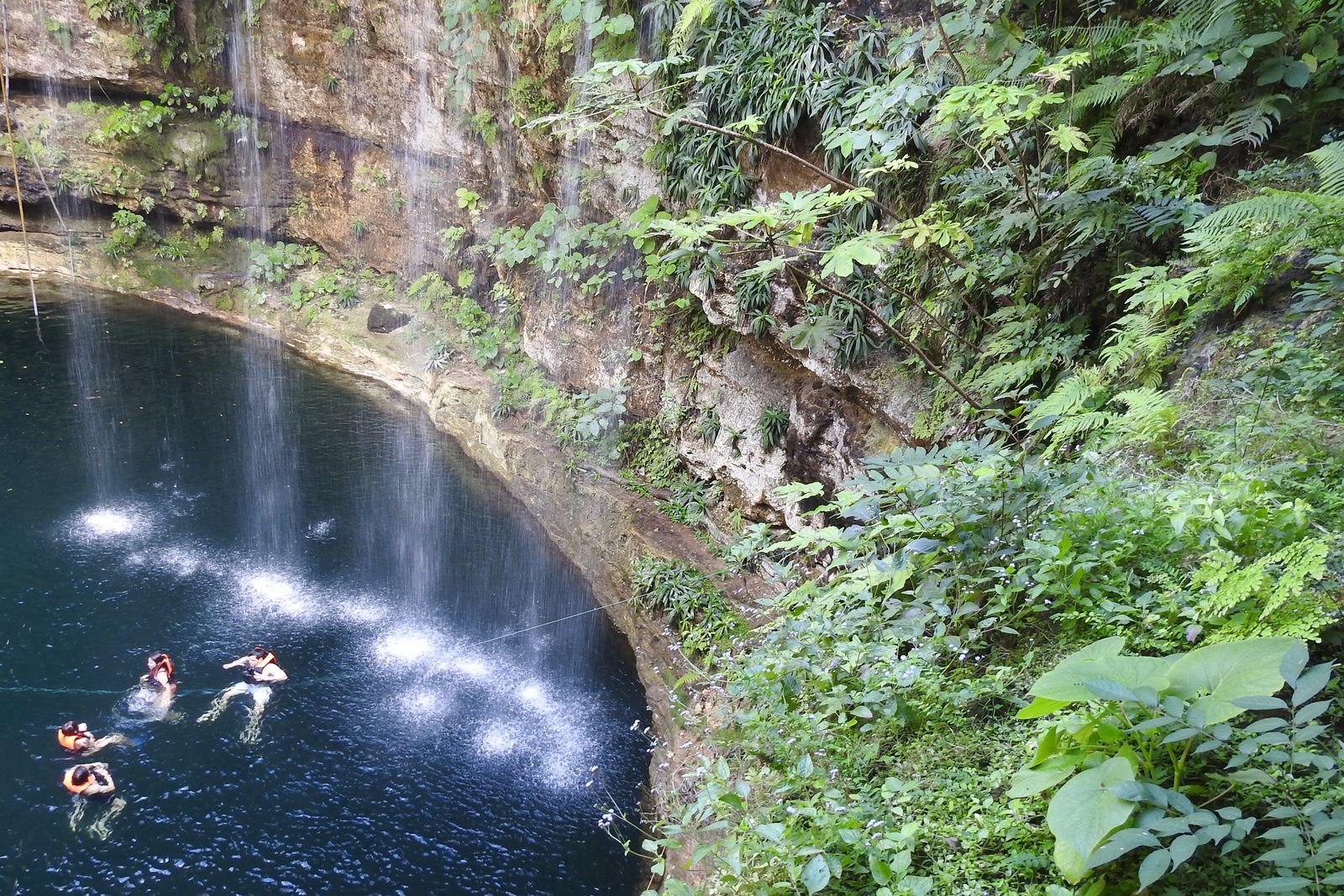 A pool of blue water at the foot of a collapsed limestone shaft, rock walls hung with vines and tree roots, swimmers near the centre