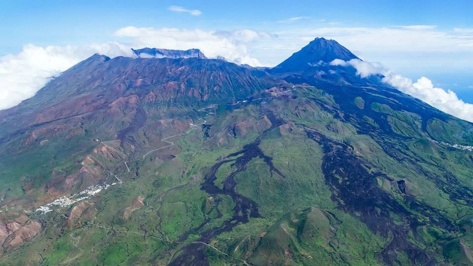 An aerial view of green slopes and dark lava flows rising to a caldera wall and a cone