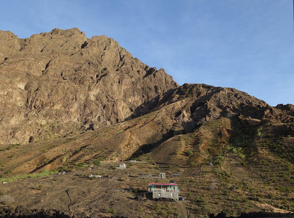 Small stone houses on dark ground below a steep caldera wall in warm light