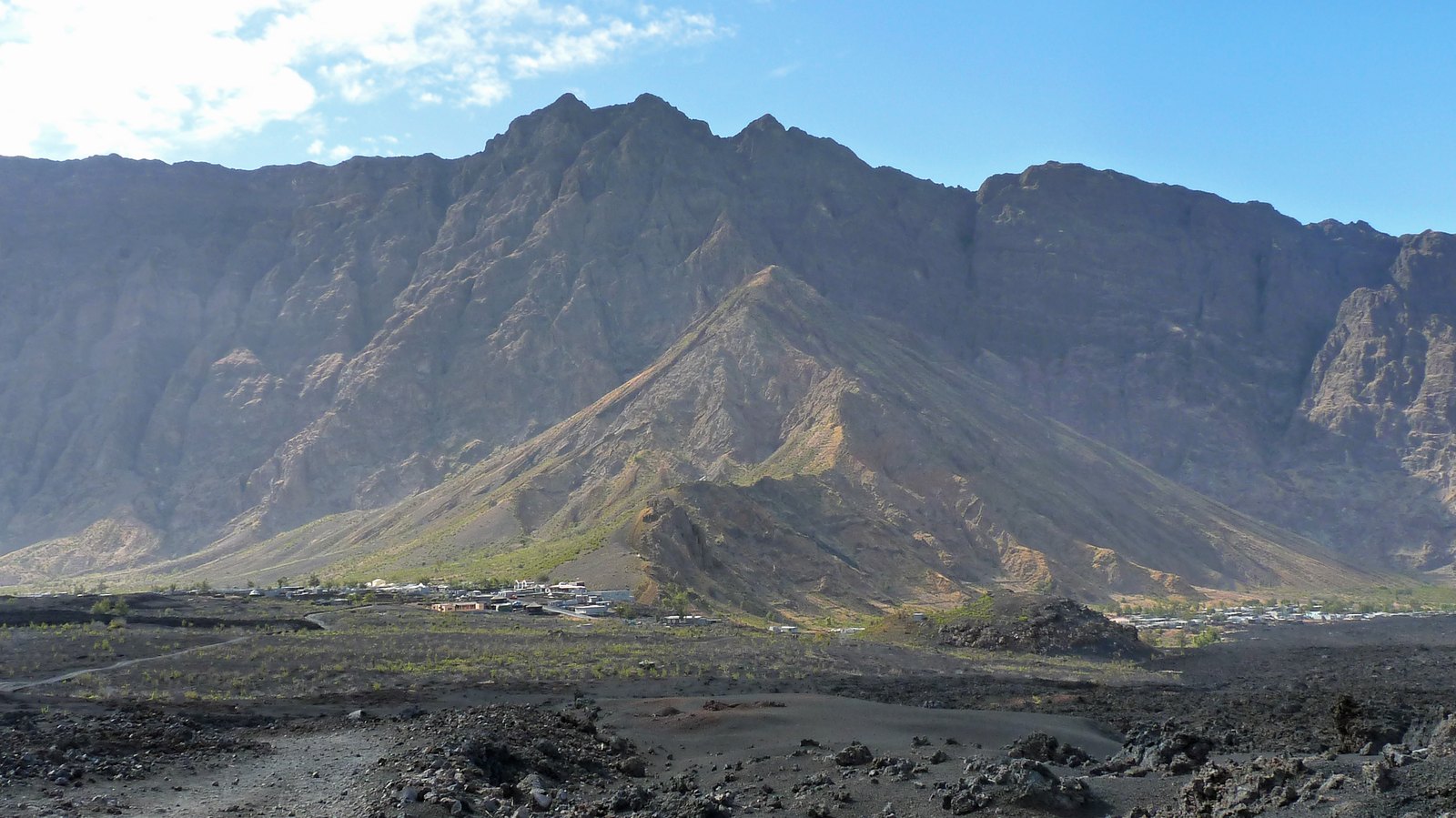 A high curved cliff wall enclosing a flat floor of black lava, two small villages of pale houses strung along its foot beneath a bare volcanic ridge