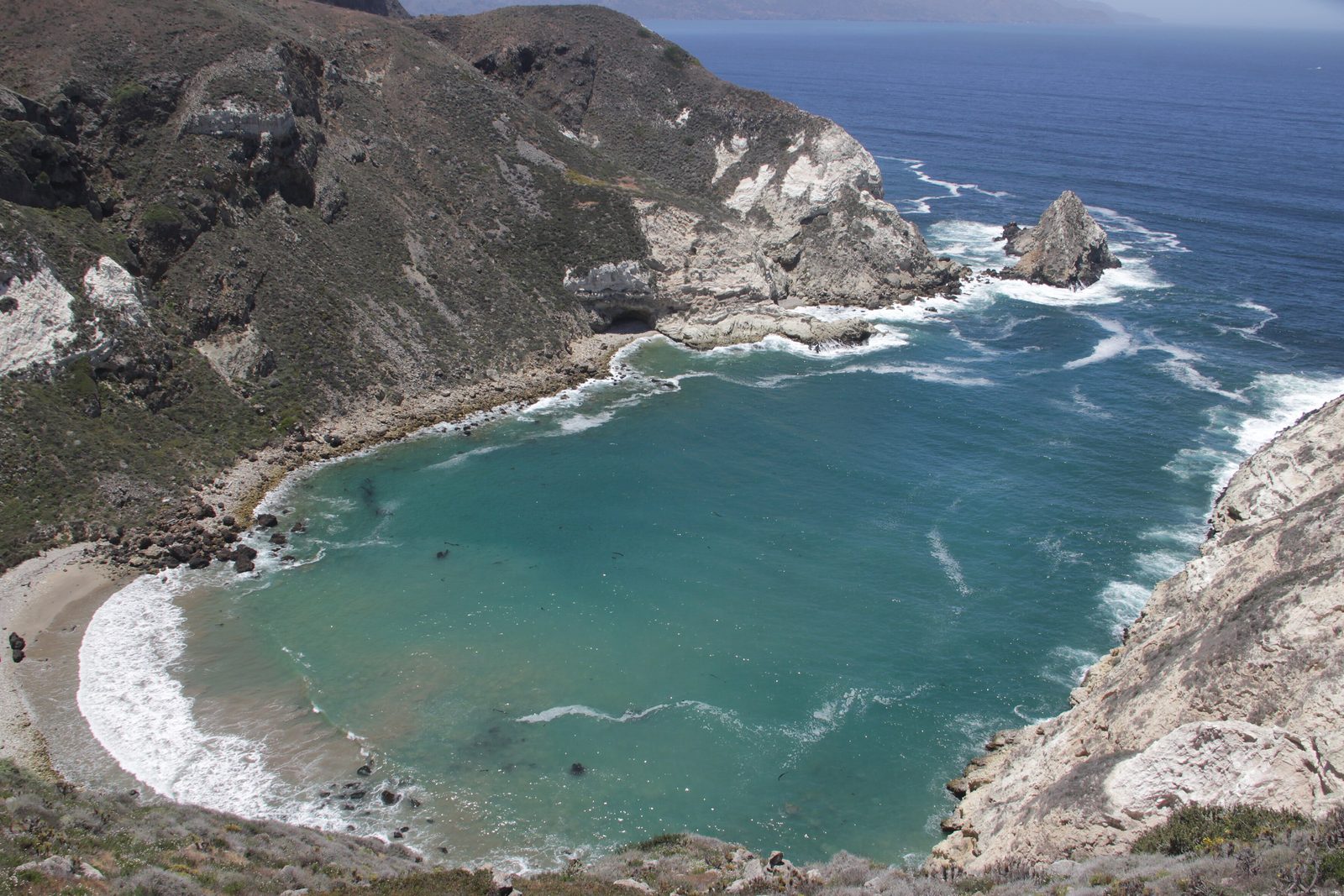 A turquoise cove ringed by steep pale cliffs and scrub-covered slopes, with open sea and a rock stack beyond