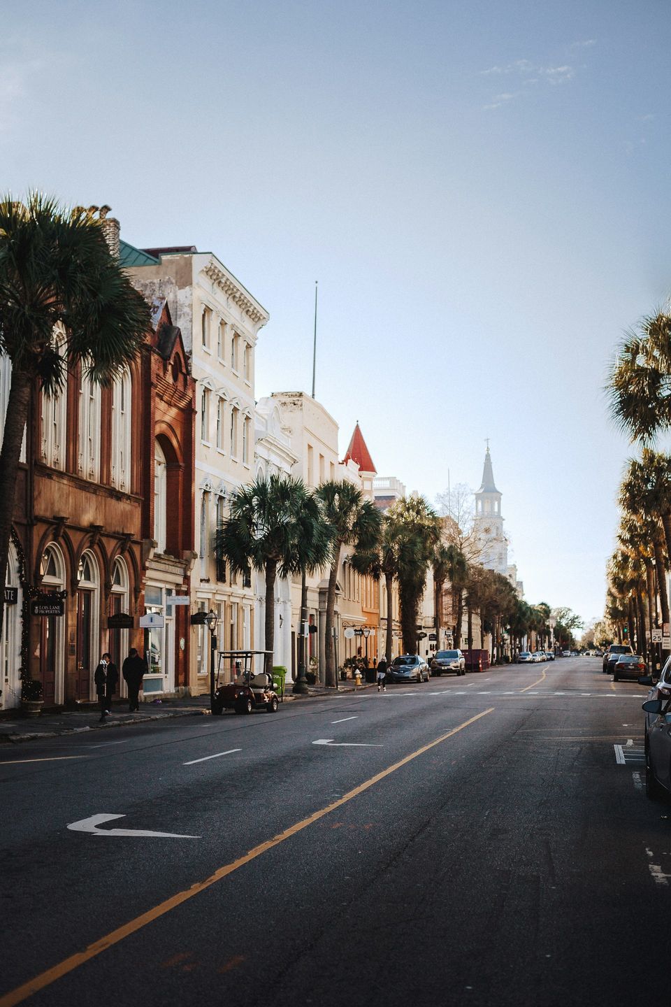 A quiet street lined with tall buildings and palm trees running toward a white church steeple