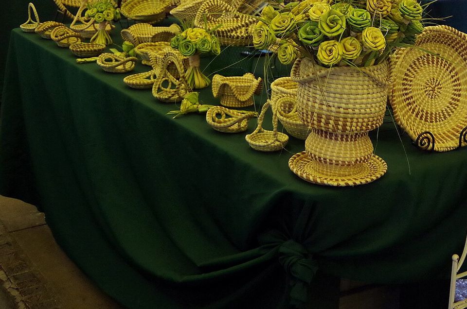 Coiled sweetgrass baskets, bowls and palmetto roses laid out on a green-draped table in a market shed