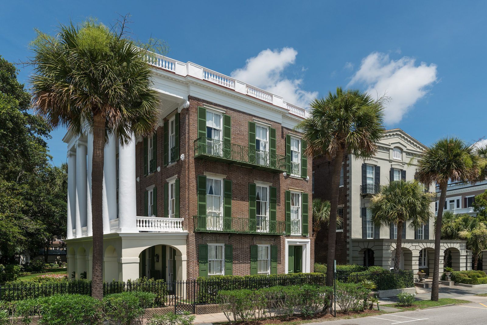 A white-columned brick mansion with green shutters beside palmetto trees on a quiet street under a blue sky