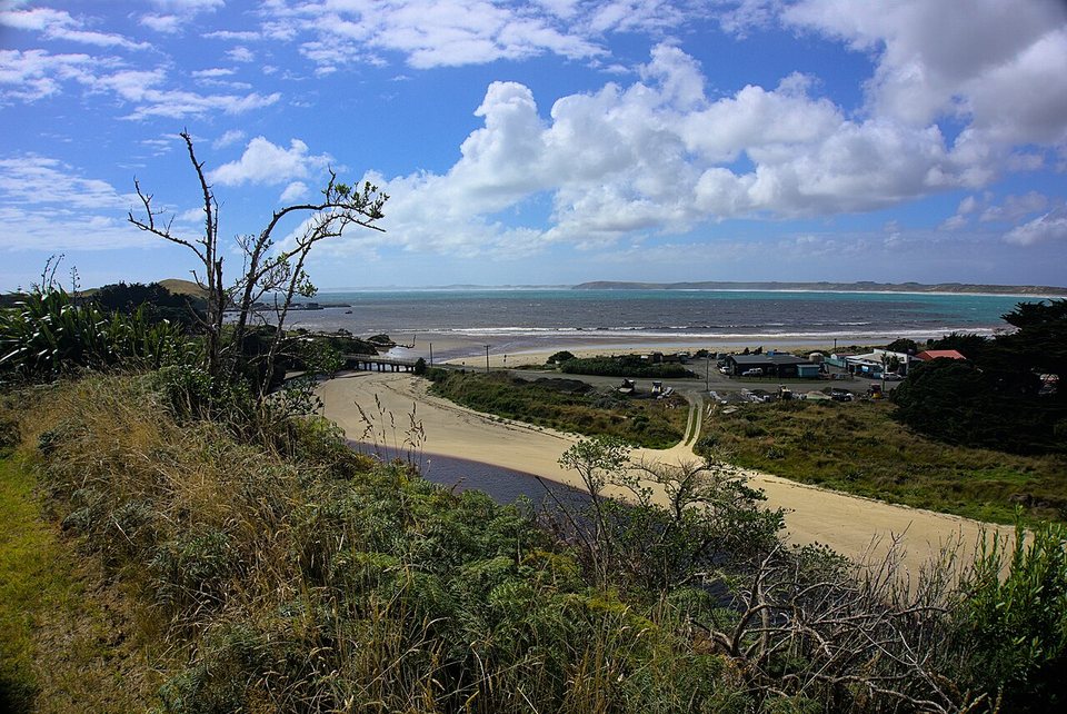 A sand track beside a bay under big clouds, with small buildings in the distance