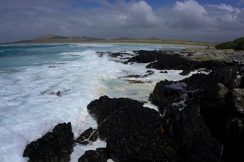 White surf on black rock beside turquoise water and a long beach under dramatic cloud