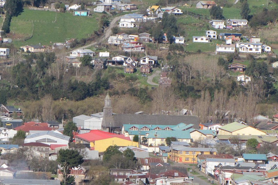 A shingled wooden church tower and long dark roof rising behind colourful painted houses on a hillside