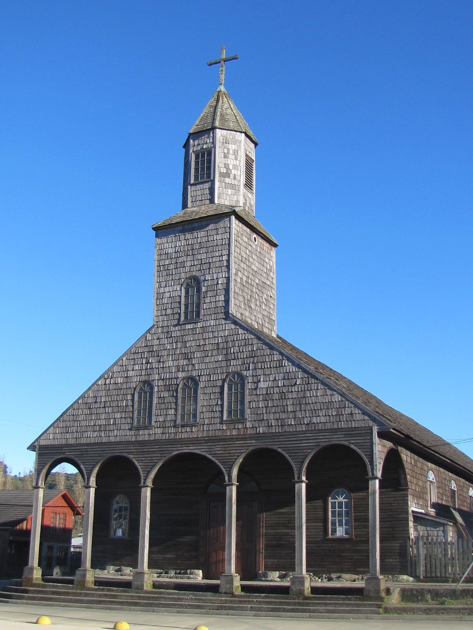 The front of a wooden church covered in grey shingles, a tiered tower above a row of arches, under a clear blue sky