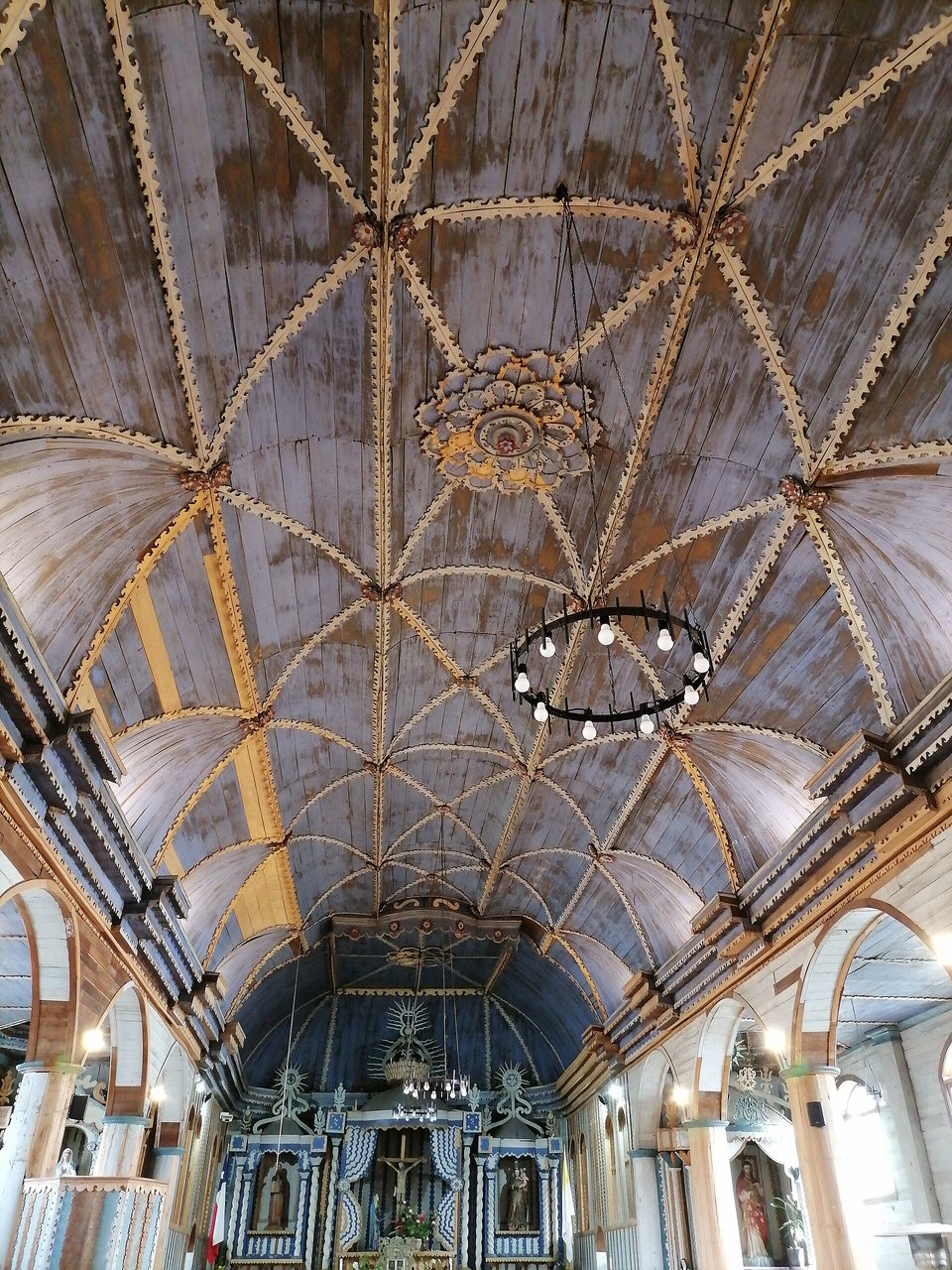 Looking up at a vaulted wooden ceiling of grey-blue boards with carved cream ribs and a central rosette, the altar small at the bottom