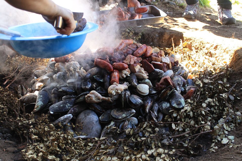 Steaming mussels, clams, sausages and meat heaped on leaves in a pit in the ground, a hand lifting food into a blue bowl