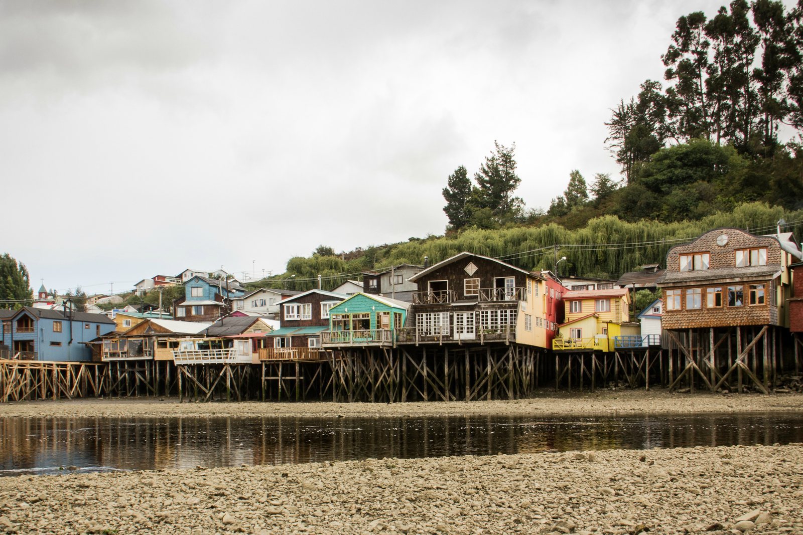 A row of wooden houses on stilts along the water's edge, their pilings exposed above the tidal flat at low water, hillside houses behind