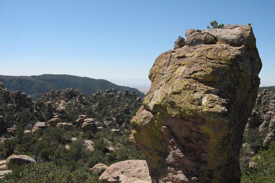 A tall lichen-covered rock pillar beside a trail, with forested ridges and rock spires behind