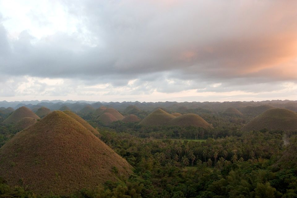 Brown grassy cone hills in dim evening light under heavy cloud with a pink glow at the right, dark forest between them