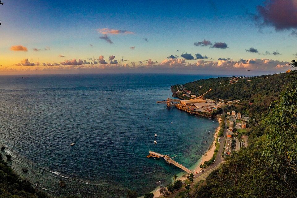 From a forested cliff, a deep-blue cove curves to a jetty, a narrow beach, a settlement and a phosphate loading works under a sunset sky