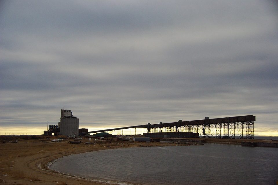 A grain elevator and a long loading gallery on a grey shore under a heavy sky, a pool of water in the foreground