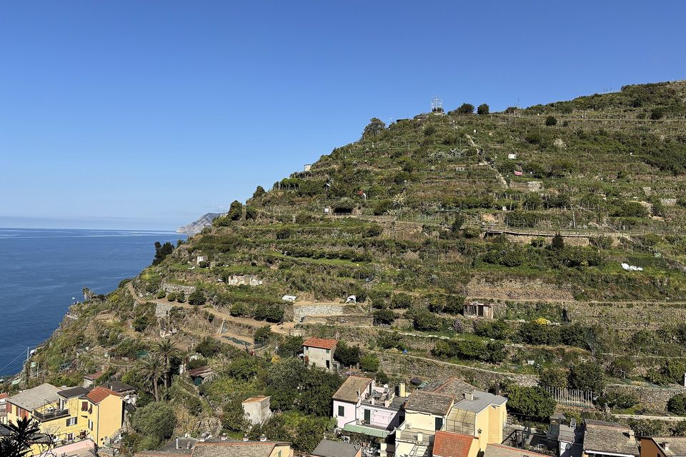 Stone-walled terraces stepping up a steep hill above a village's roofs, the sea at left under a clear sky
