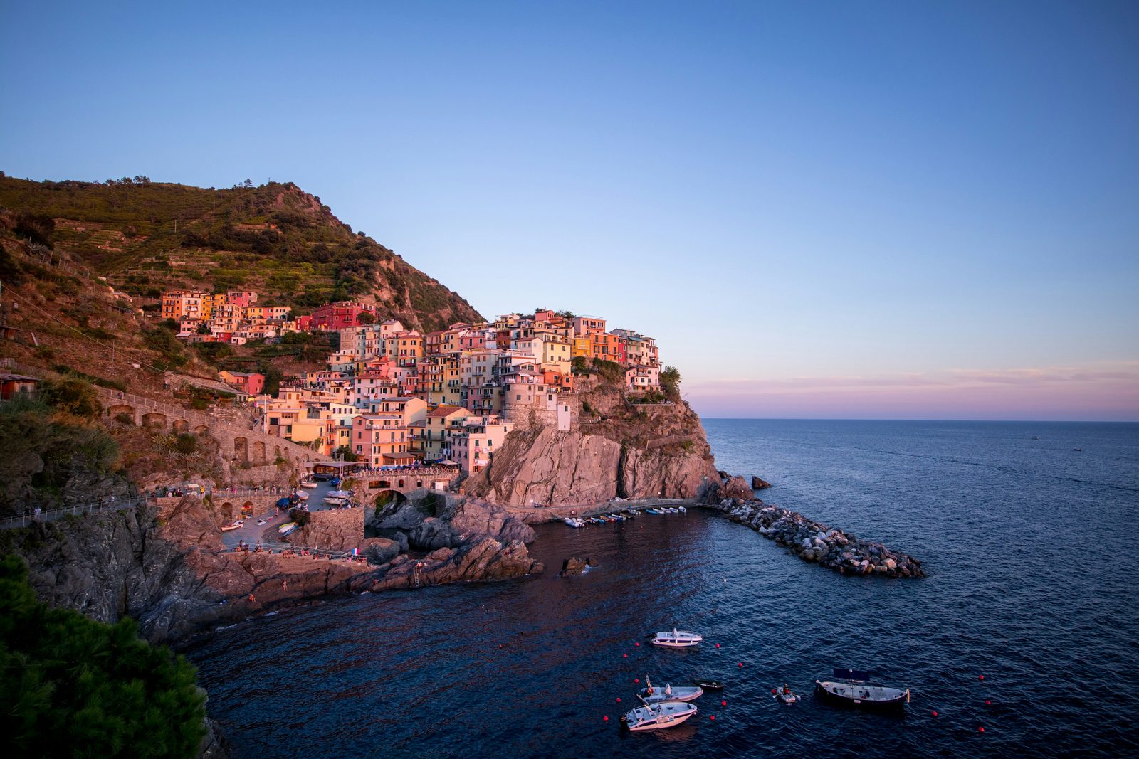 Colourful houses stacked on a rock above a small harbour in soft pink light, a terraced hill behind