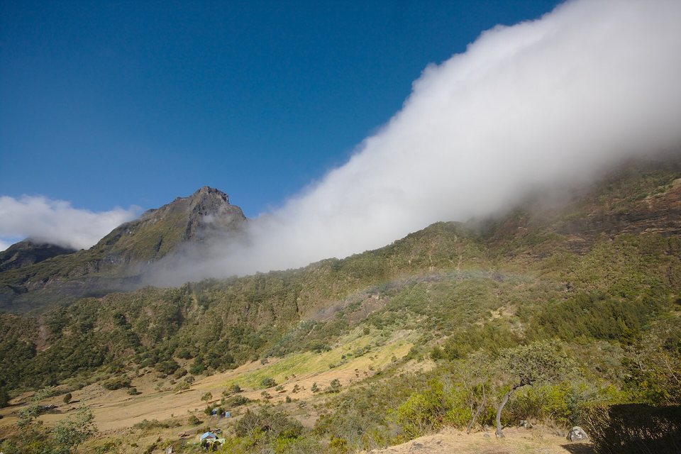 A bank of white cloud spilling over a green mountain wall under a deep blue sky, a grassy plain with a few small huts below