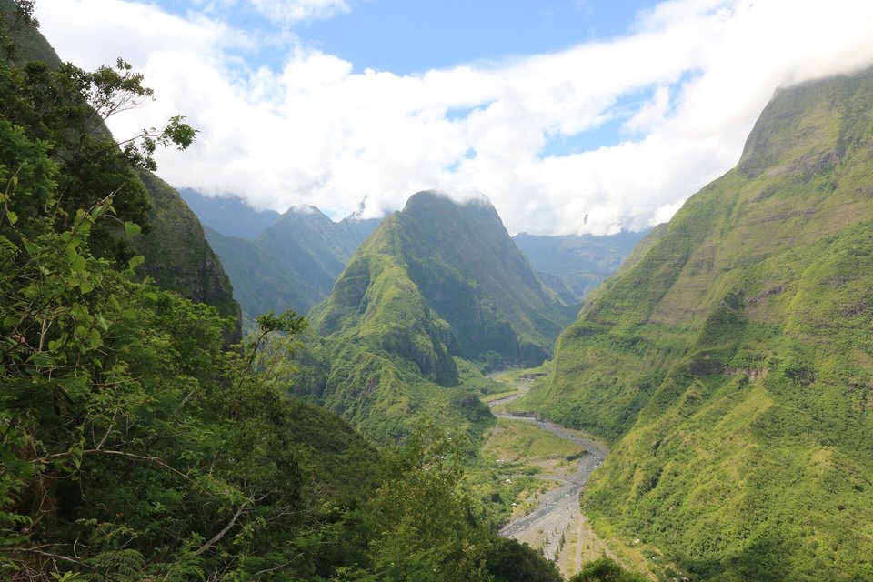 A grey stony riverbed winding between steep green ridges into a valley of pointed peaks under cloud