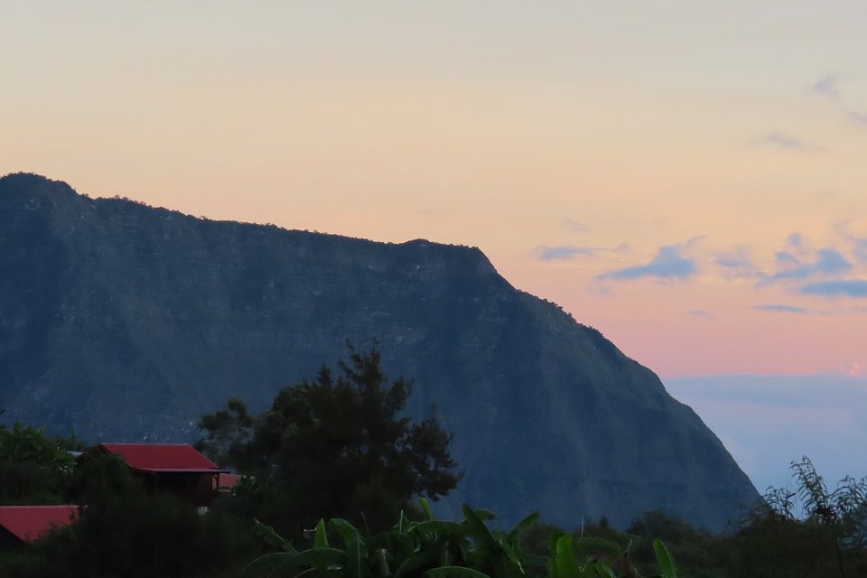 The dark silhouette of a long flat-topped mountain wall against a peach and pink dawn sky, red roofs in the foreground