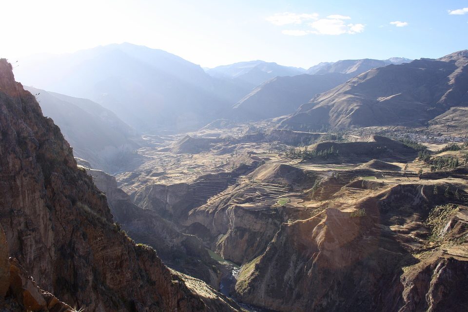 A dark rock wall at the left above a deep gorge, terraced benches and distant ridges in hazy backlight