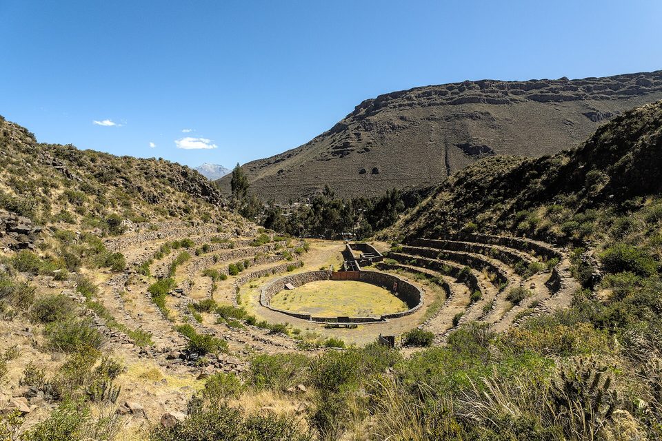A circular grassy terrace ringed by curved dry-stone walls stepping up a dry hillside, a bare ridge behind under a blue sky