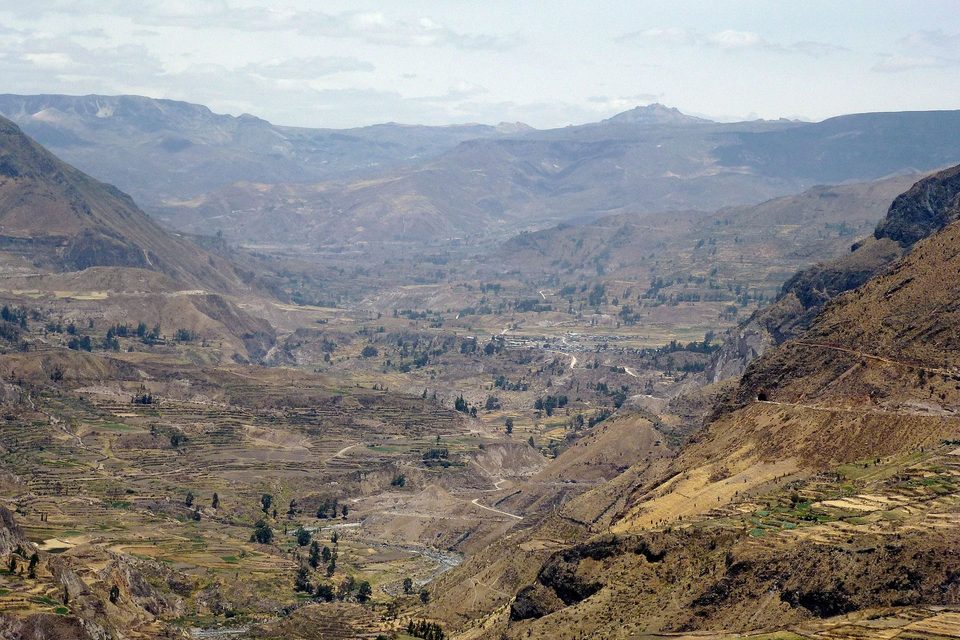 A wide hazy valley with terraced slopes stepping down to a small river and a distant village, ridges beyond