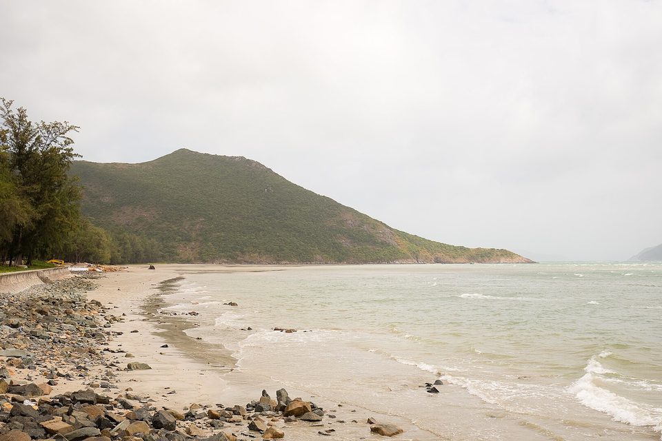 A curving sand-and-pebble beach meets a pale-green sea below a forested headland, under grey cloud