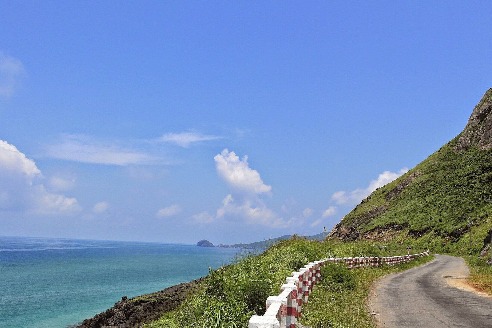 A narrow coast road with a red-and-white railing curves along a green, rock-faced hillside above turquoise water, with small islets on the horizon under a bright blue sky